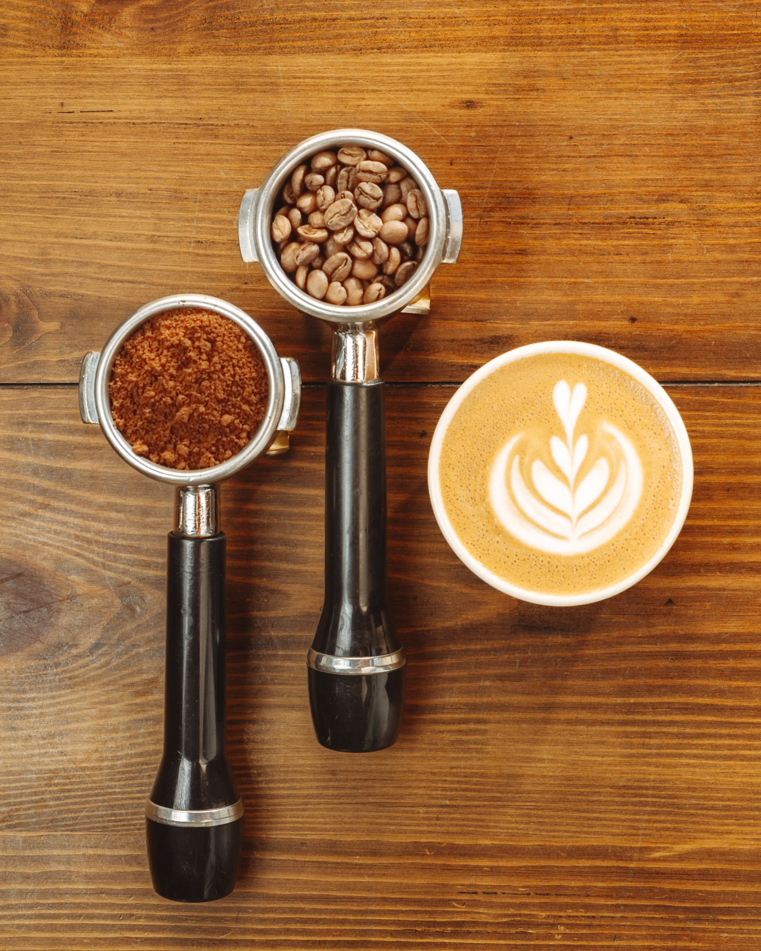 Two portafilters with ground coffee and beans beside a latte